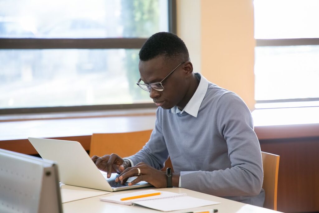 Young black man working on a laptop near a window in a library